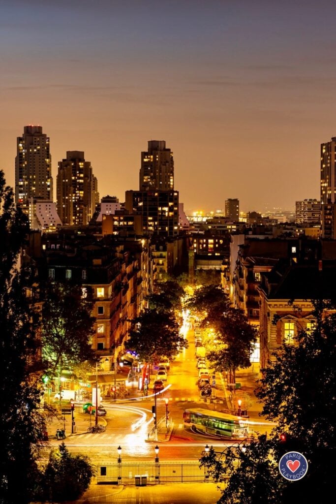 Elevated view down illuminated Parisian street at night with golden street lights creating light trails, flanked by residential buildings and modern towers against orange sunset sky