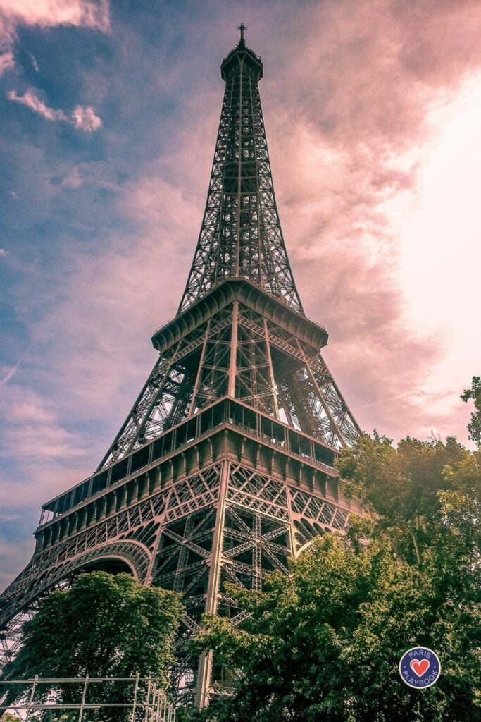 Low-angle view of Eiffel Tower rising through green tree canopy against dramatic pink and purple cloudy sunset sky showing intricate iron lattice work and architectural details