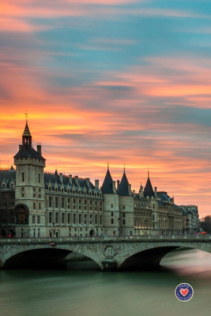 Conciergerie palace and medieval towers at sunset with dramatic orange and turquoise sky reflecting on Seine River with stone bridge in foreground