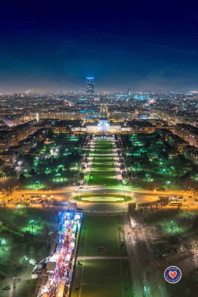 Aerial night view of Champ de Mars park with illuminated green lawns leading to École Militaire, Christmas market lights, and Montparnasse Tower against deep blue sky