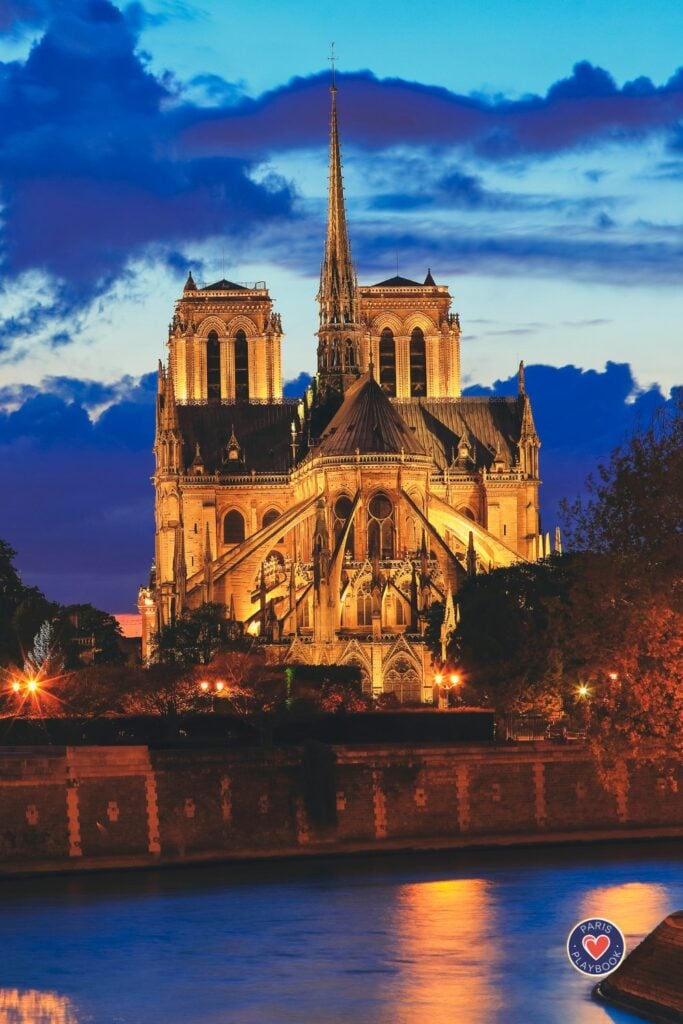 Notre Dame Cathedral illuminated in golden light at blue hour with twin towers, spire, and flying buttresses reflected in Seine River against dramatic cloudy sky