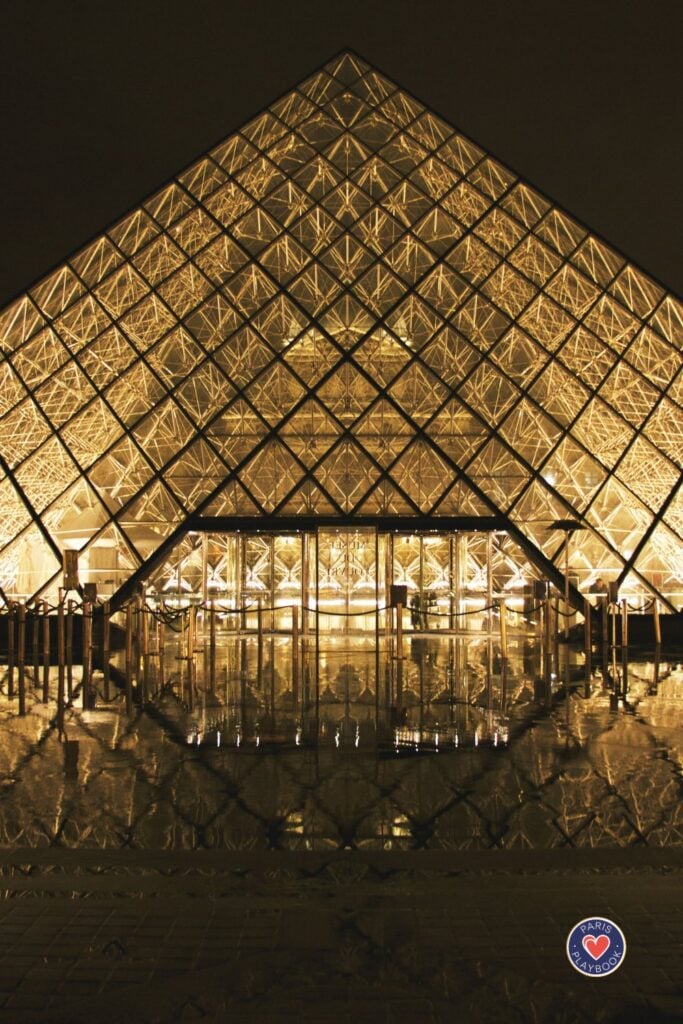 Louvre Pyramid illuminated at night with geometric glass panels creating diamond patterns and golden reflections with visitors silhouetted beneath the glowing structure