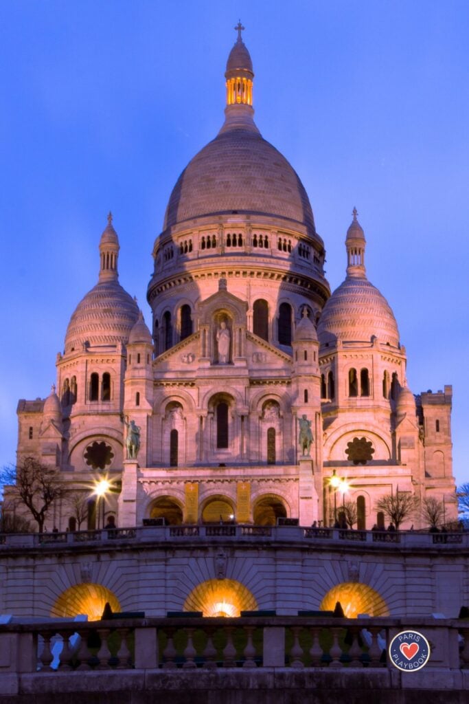 Sacré-Cœur Basilica illuminated in warm golden light at blue hour with white stone domes and Byzantine architecture against purple twilight sky with illuminated arches in foreground