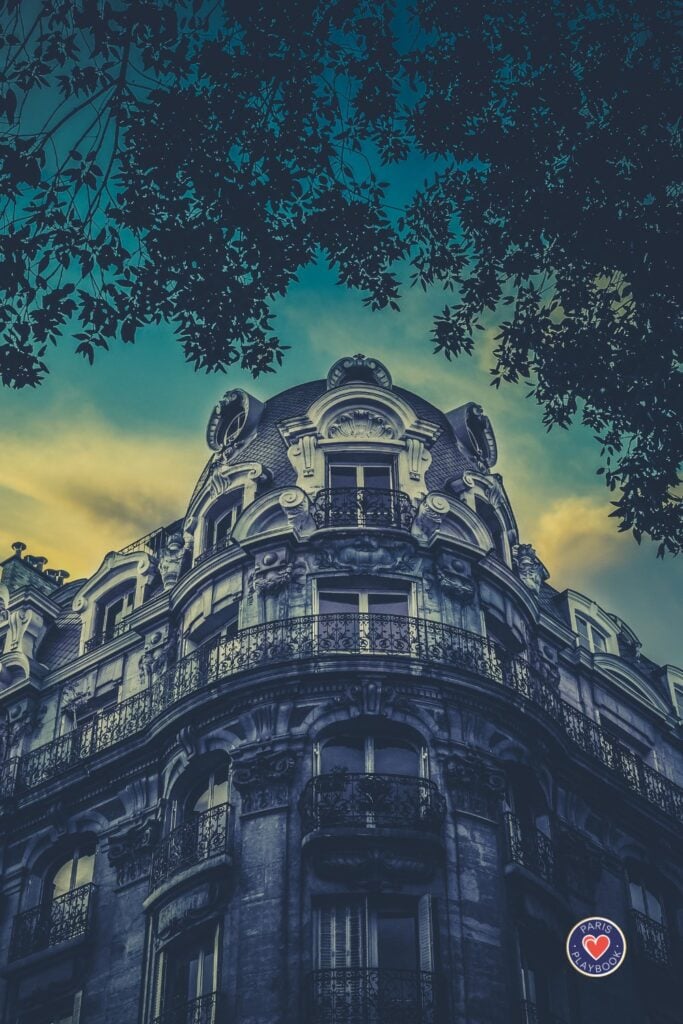 Ornate Haussmann-era Parisian building with elaborate balconies and architectural details silhouetted against dramatic teal and yellow sunset sky with tree branches framing the scene