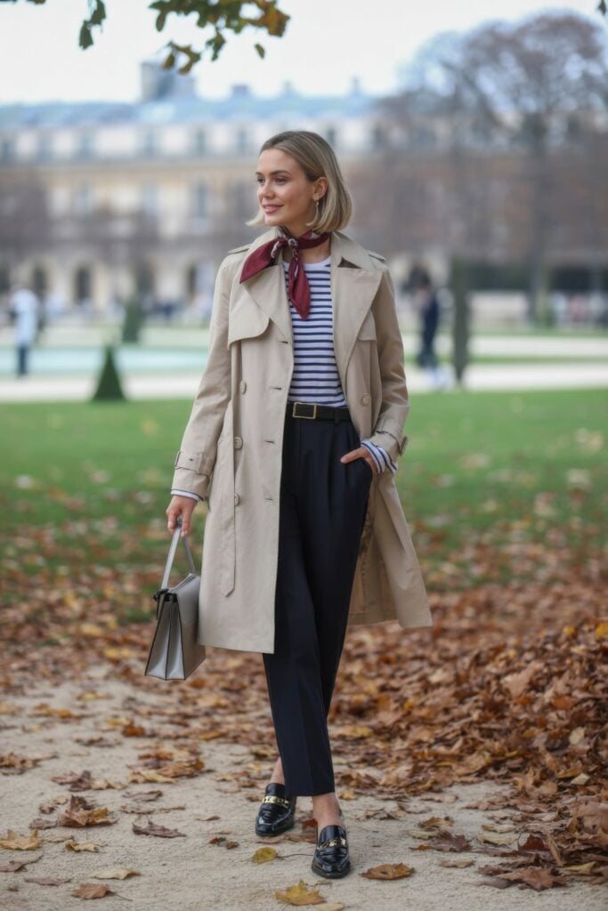 Woman wearing beige trench coat over striped shirt with silk scarf, black trousers, and loafers in Parisian park