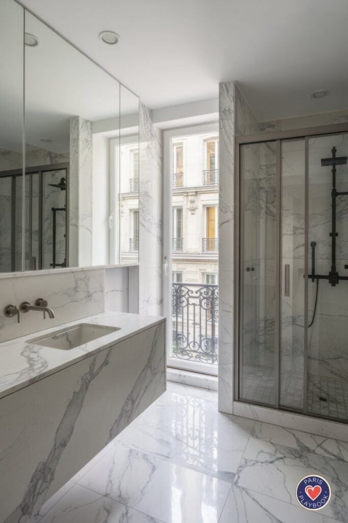 Minimalist Parisian bathroom with floor-to-ceiling white marble, black-framed shower enclosure, floating marble vanity, and French doors opening to wrought-iron balcony