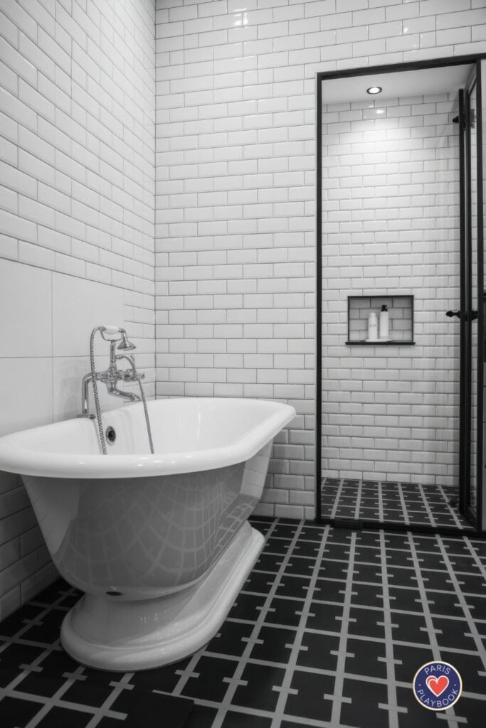 Black and white Parisian bathroom with white subway tiles, black geometric cross-pattern floor tiles, white pedestal tub, and black-framed glass shower enclosure