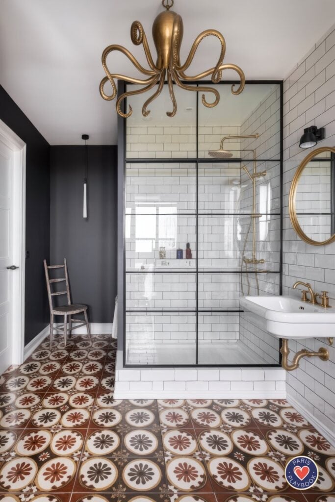 Eclectic Parisian bathroom with brass octopus chandelier, charcoal gray walls, white subway tile shower with brass fixtures, round brass mirror, patterned Moroccan floor tiles, and black-framed glass shower enclosure