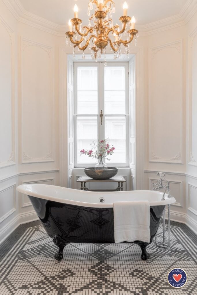 Elegant Parisian bathroom with glossy black and white clawfoot tub, intricate mosaic tile floor medallion, white wall paneling, gold crystal chandelier, tall French windows, and antique side table